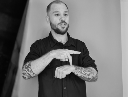 Foto preto e branca de Giliard sinalizando em Libras em Estúdio. Giliard é homem branco, de cabelos e barba loiras. Veste uma camisa preta de botão.