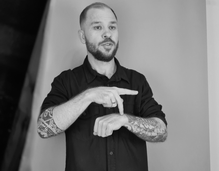 Foto preto e branca de Giliard sinalizando em Libras em Estúdio. Giliard é homem branco, de cabelos e barba loiras. Veste uma camisa preta de botão.